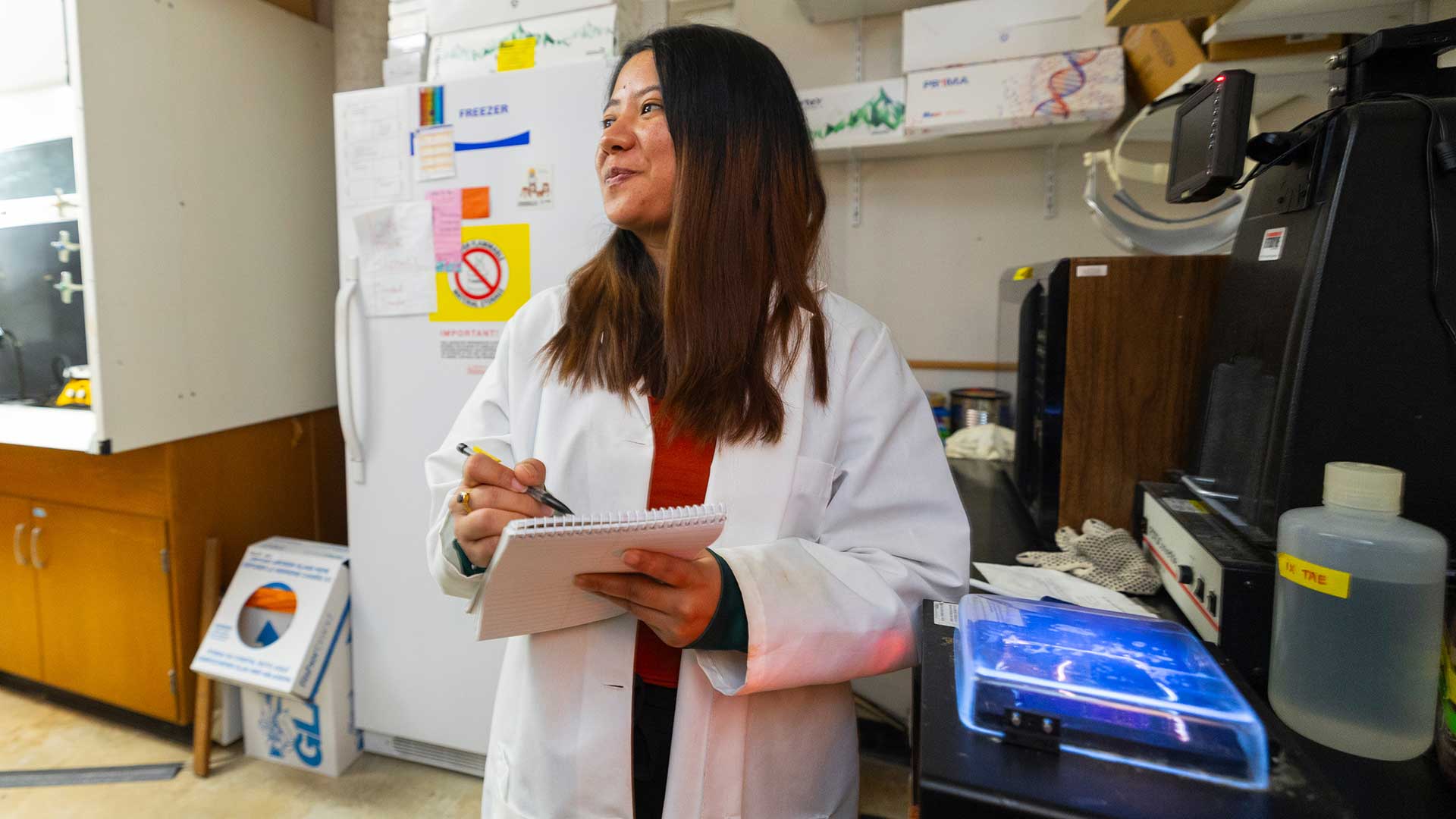 A graduate researcher smiles while working in a lab and holding her notebook.